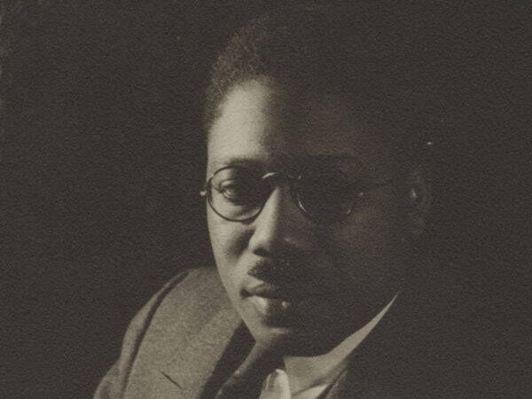 Black and white photo of African American man with close-cropped hair, wearing glasses, and a suit, against a dark background, with light comeing from the left of the photo. There is a sliver of a print curtain at the left of the image.