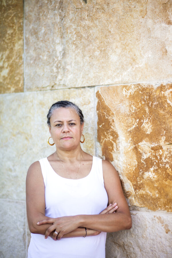 Photo of a black woman wearing a sleeveless white shirt, gold necklace, silver rings and gold hoop earrings standing in front of a brown and ivory limestone wall