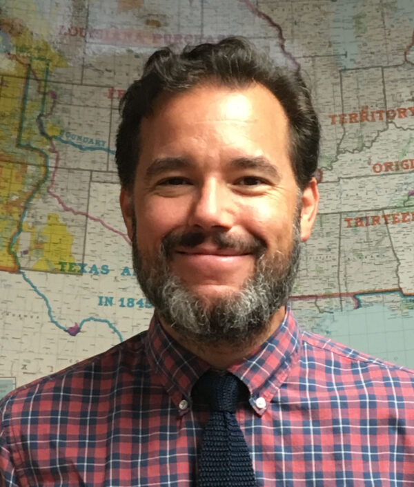 Head and shoulders photo of a man with dark, curly hair and a salt-and-pepper beard, standing in front of a map. He is wearing a red and blue small plaid pattern shirt, with a button down collar and a solid, dark tie