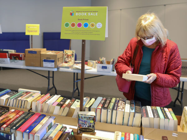 White woman with blonde hair, red jacket and mask looks at a book while standing over a table full of dozens of books with a table behind her stacked with used books