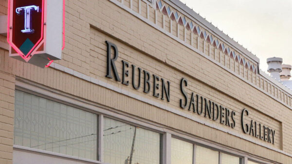 Top of a storefront, light brick, with words Reuben Sanders Gallery inset in brown. At far left is tip of a neon sign with the letter T in white on a dark purple background outlined in red.
