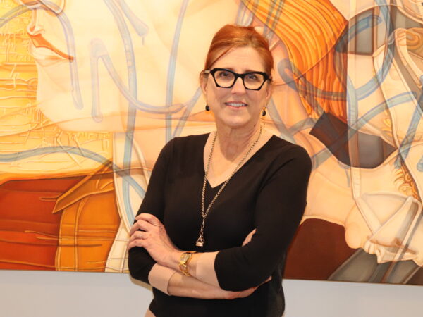 Red-haired woman with dark-framed glasses, dressed in a black v-neck with a long gold necklace, standing in front of an abstract art in yellow and orange tones