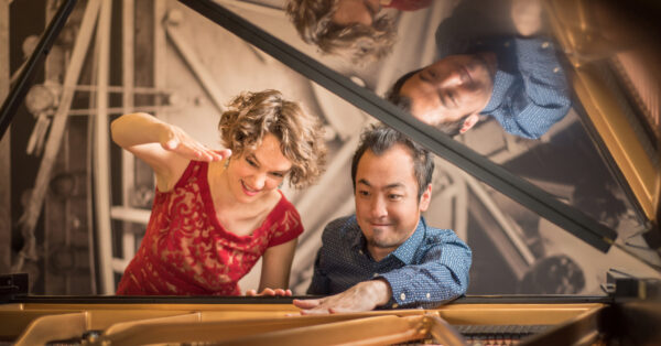 Woman and man framed by the lifted cover of a grand piano. Both are looking down, woman has an arm raised. She is dressed in a red sleeveless top and has light brown curly hair. The man, on the right, has one hand on top of the piano and is wearing a long-sleeved blue shirt