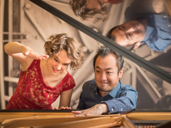 Woman and man framed by the lifted cover of a grand piano. Both are looking down, woman has an arm raised. She is dressed in a red sleeveless top and has light brown curly hair. The man, on the right, has one hand on top of the piano and is wearing a long-sleeved blue shirt