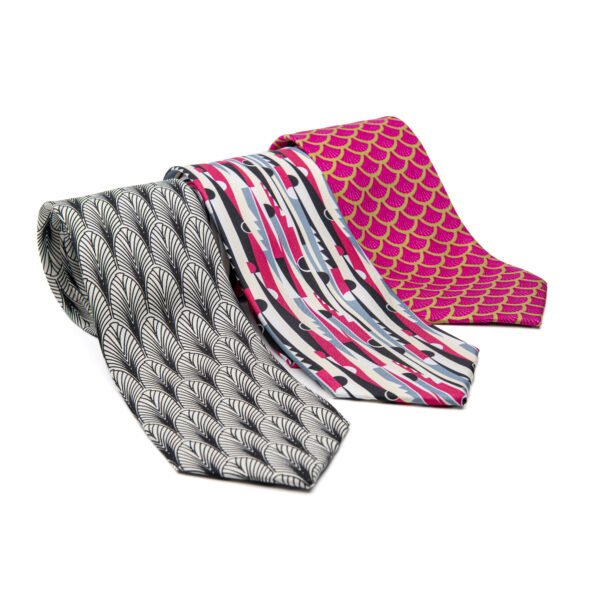 Three men's ties, rolled, side-by-side. Far left is black and white geometic pattern. Center is brightly colored reds blues, and blacks in a striped pattern, right is red and gold in an art-deco-inspired fish scale pattern