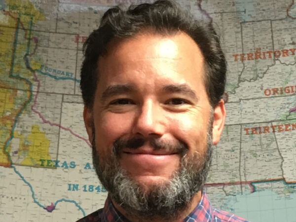 Photo of a white man with dark hair , dark moustache and salt-and-pepper beard standing in front of a map of the United States