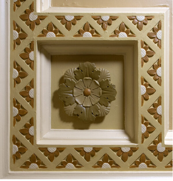 Round flower carving in shades of brown, in a square frame against a background with a floral motif in shades of brown.