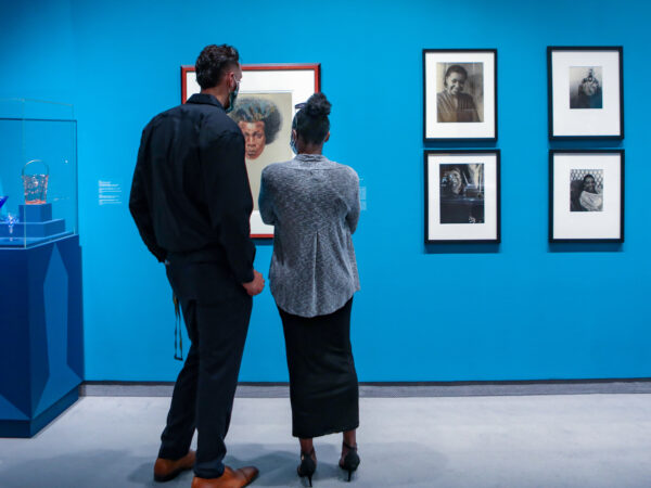 Man and woman standing in gallery in front on a painting of an African American man. On their left is a pedestal with an enclosed top an several pieces of glass. On their right is a collection of six photographs. Gallery walls are painted a bright blue. and six photographs