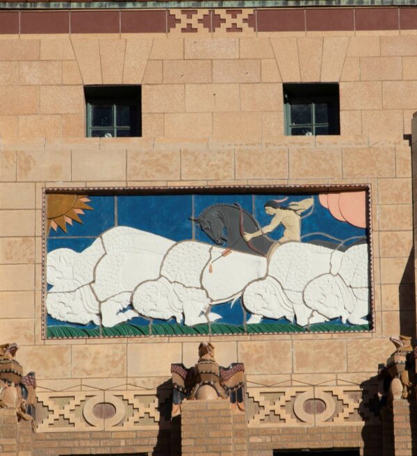 Relieft sculpture depicting white buffalo hunted by an Indian on a black horse against a blue sky, with an orange and yellow sun in the top left corner and a pink cloud in the top right corner. Below the mural are three eagle sculptures in brown tones