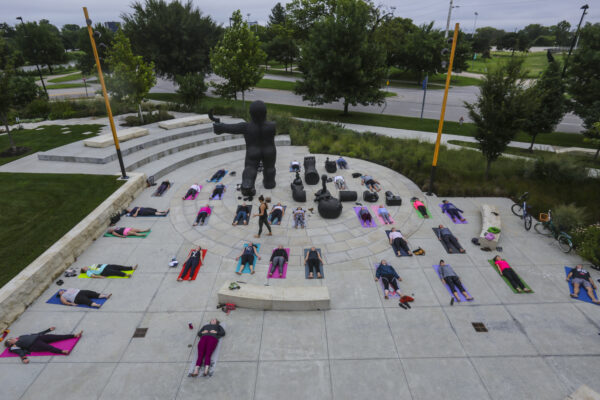 Exterior photo of the Jayne Milburn Sculpture Plaza where nearly two-dozen people are laying on the concrete plaza on colorful mats on their backs in a yoga pose
