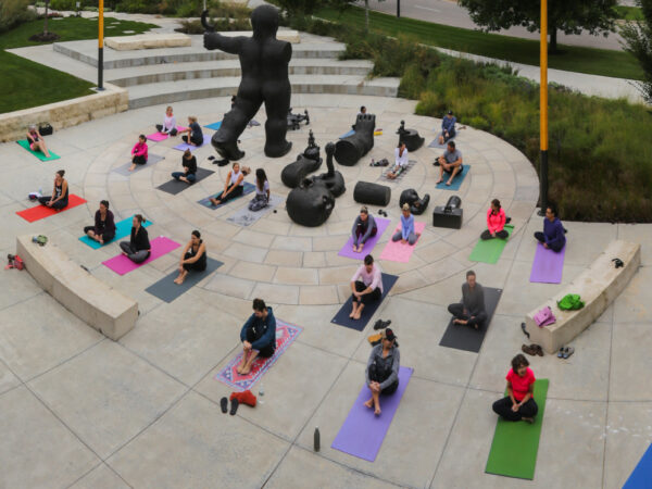 Exterior photo of the Jayne Milburn Sculpture Plaza where nearly two-dozen people are laying on the concrete plaza on colorful mats sit cross-legged in a yoga pose