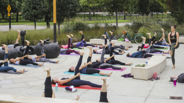 Exterior photo of the Jayne Milburn Sculpture Plaza where nearly two-dozen people are laying on the concrete plaza on colorful mats with their left legs stretched up in the air in a yoga pose