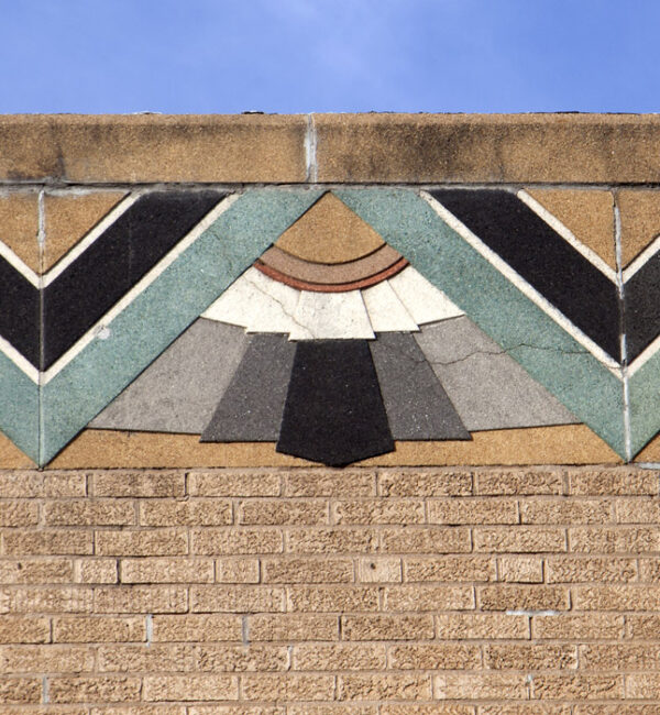 Building facade of buff brick with top decoration of turquoise, black and gray-toned stone. Center is a half-circle of three layers of brown, with white.