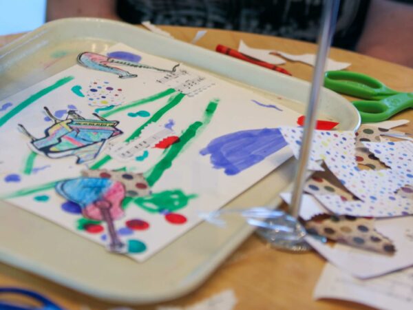 Colorful abstract drawing with green lines, purple and blue shapes, on an off-white tray with pencils, other art supplies, and other drawings on the tabletop around it.
