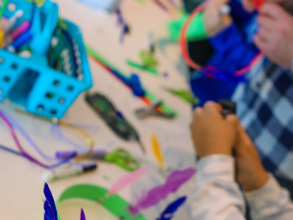 A child's and an adult's hands making art with strips of colored paper next to a container of scissors on a white tabletop