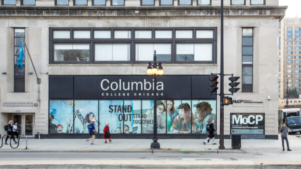 Street scene with stone building facade. Sign in the center over a series of windows identifies building as Columbia College Chicago. Underneath the sign in the windows is a collage of young people and text Stand Out, Stand Together. At the far right at street level is a sign, white on black, MoCP in large letters over Museum of Contemporary Photography in smaller letters. At the far left is two people walking toward to right, In the center are two more people on sidewalk. Center right are two more people walking on the sidewalk and in the right corner is a man standing at the corner facing to cross the street.