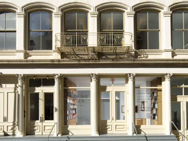 Two stories of a building on white stone. Gray steops span the width of the building on the street level. Five columns are across the first level, with wood and glass doors. The second level has recessed window with carved arches and an metal balcony with round scrolls. Doors in th center ar flanked with tall windows with writing. On the left, the window says The Drawing Center.