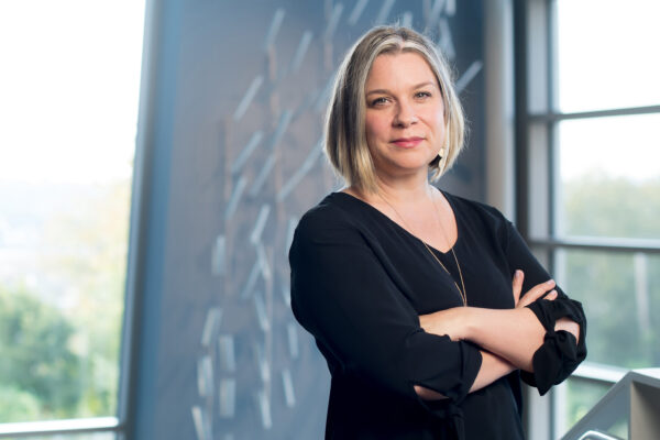 White woman standing with arms crossed. She has medium length blonde hair and is dressed in black with a gold necklace. She stands in front of a window and the wall behind her has a reflection of a geometic pattern.