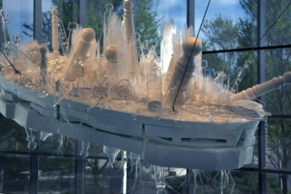 Hanging sculpture constructed of clear glass objects like vases, bowls and white poles resembing barber poles on a white base with clear glass objects resembling roots on the underside of the white base. Black cables holding the sculpture are visible in the right side of the image. Behind the sculpture are floor-to-ceiling windows looking out on treetops covered in green leaves.