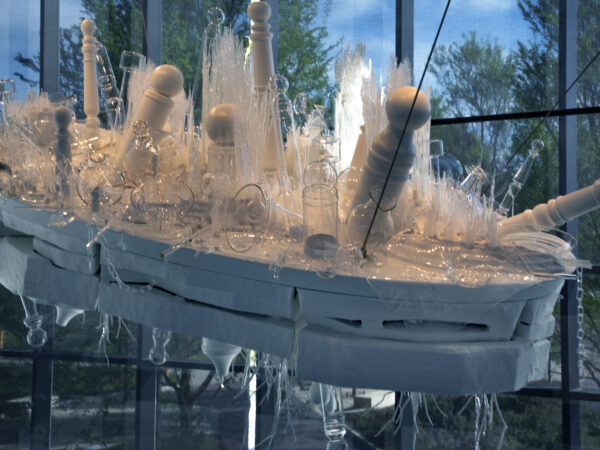 Hanging sculpture constructed of clear glass objects like vases, bowls and white poles resembing barber poles on a white base with clear glass objects resembling roots on the underside of the white base. Black cables holding the sculpture are visible in the right side of the image. Behind the sculpture are floor-to-ceiling windows looking out on treetops covered in green leaves.