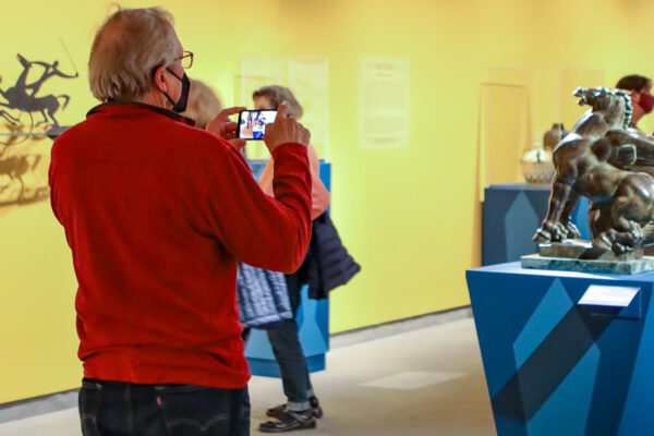 Photo of a white man with grey hair, glasses, a mask, red sweater and dark pants uses his smartphone in the galleries to photograph a bronze horse sculpture on a blue pedestal with three museum visitors in the background