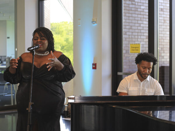 Black woman dressed in an off the shoulder black gown with a wihite necklace, in front of a microphone, her hands outsteteched. To her left is a black man dressed in a white shirt, playing a black grend piano.