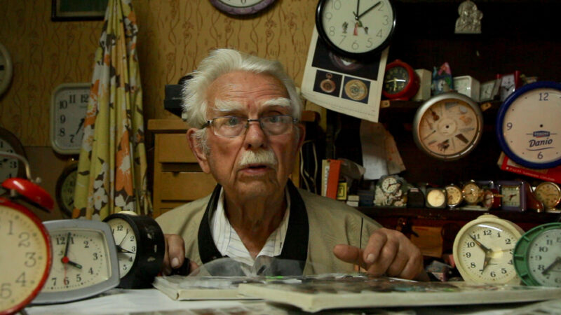 Head and shoulders image of older white man, with white hair and mustache, wearing a tan jacket and white shirt, left hand pointing to an open book on the table in front of him and surrounded by various clocks.