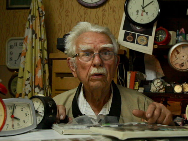 Head and shoulders image of older white man, with white hair and mustache, wearing a tan jacket and white shirt, left hand pointing to an open book on the table in front of him and surrounded by various clocks.