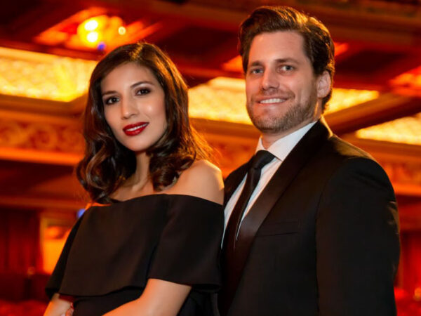 Couple, both dressed in black against an background of an opulant ceiling in reds and golds
