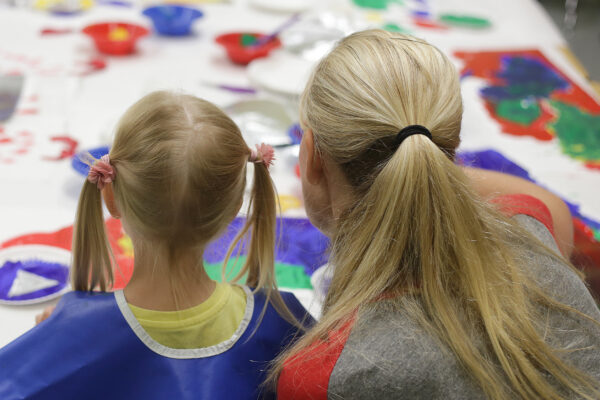 Blonde girl with pigtails, wearing a blue top and woman with blonde hair, pulled into a ponytail, wearing a gray top with red sleeves, seen from the back, painting a car in bright blue, red, and green on white paper.