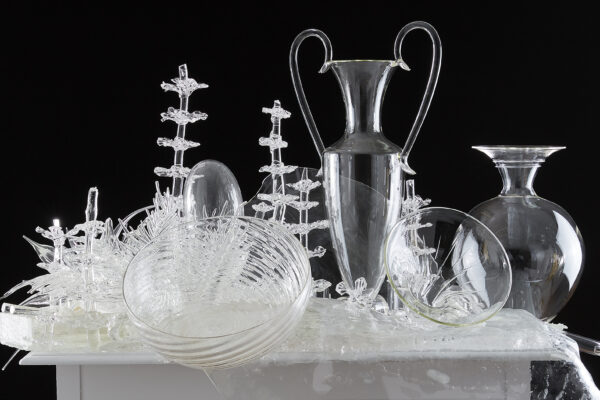 White tabletop covered in artworks of clear glass, including two vases, two bowls, and plant-looking forms