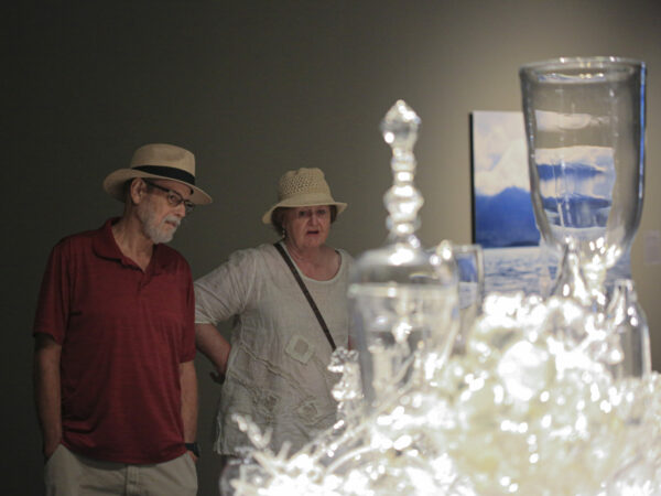 People looking at a glass sculpture featured vases, bowls, jars and forms. Man at left wears a wide-brimmed straw hat and a red shirt. Woman at right wears a wide-brimmed straw hat andis dressed in a white shirt with floral print and has a black purse strap across her torso.