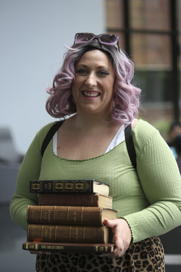 Woman with blonde, shoulder length hair, with sunglasses pushed to the top of her head, dressed in a light green shirt, carrying a pile of 5 books.