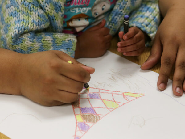 A Black mother's hands hold a yellow crayon while her child's hand holds a purple crayon while they color a party hat on a white sheet of paper