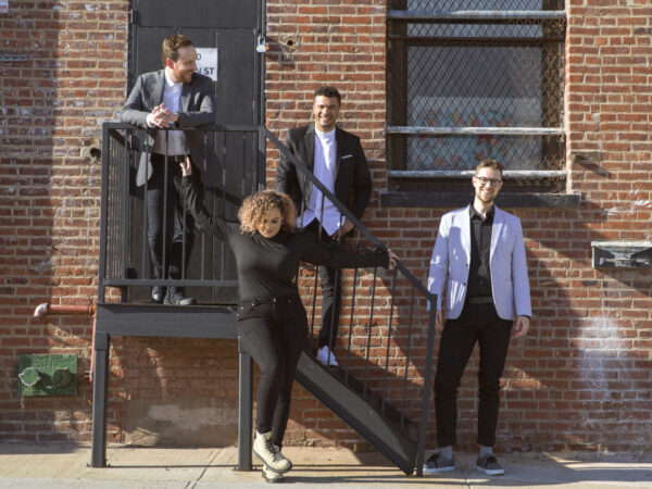 Photograph of four people standing on or in front of a metal staircase on the side of brick wall with a door at the top of the staircase on thr left and a window on the right. From left, there is a man earing a dark suit and white shirt standing at the top of the srairway, standing two steps down is a white man earing a dark suit untucked white shirt. Standing at the bottom of the stairway is a white man weaing a white jacket, dark red shirt and dark slacks. IN fron the stairway is a woman with dark, curly hair dressed in black, She is looking down and her arms are outstretched, resting on the stair rail.