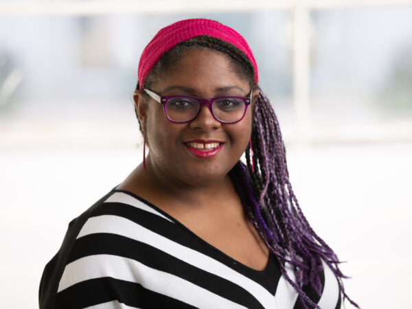 Black woman facing the camera and smiling, , with a red headband, and dark-framed glasses. Dressed in a v-neck black and white stroped shirt