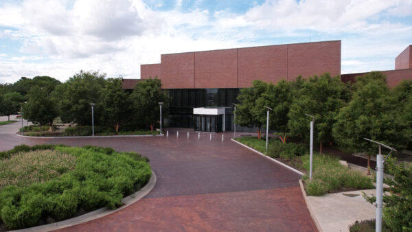 Photo of the museum's front entrance with green trees on either side showing the red, brick building, floor to ceiling windows, and the red brick Lattner Walker Family Plaza that creates a circle drive