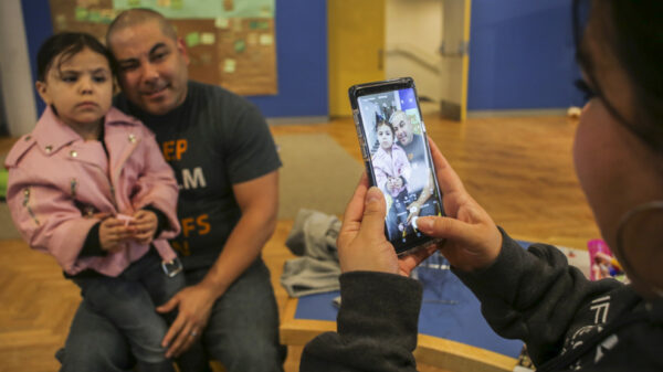 Photo of a woman on the right with dark hair and a dark grey sweatshirt holding a cell phone to take photo of a man in a short-sleeve shirt and jeans holding a young girl with dark brown hair wearing a pink jancket