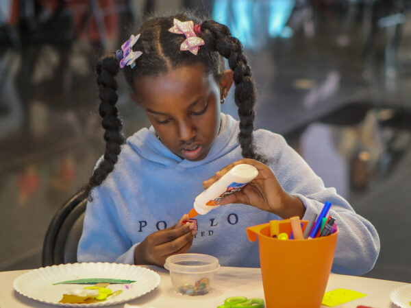 Young African American girl, her hair in two braids and wearing a purple, long-sleeved shirt. She's sitting at a table, holding a bottle of glue in her hand and applying glue to an object in her other hand. Various art supplies are on the table front of her.