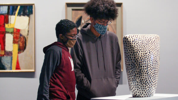 Two young black men wearing masks and sweatshirts look at a white sculpture with black dots on a pedestal in the middle of the Wichita Art Museum galleries