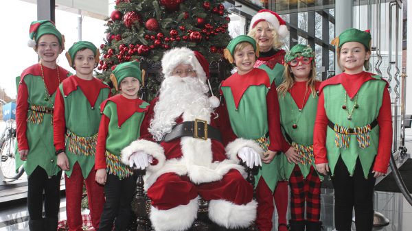 Santa Claus sitting on chair in front of a Christmas tree with Mrs. Claus behind him and five young people dressed in red and green elf costumes