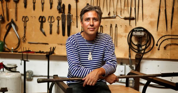 Mand dressed in black and white striped shirt sitting in a workshop with tools on a peg board all behind him.