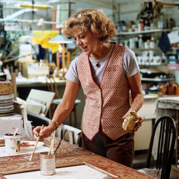 Woman standing at a table, her right hand oustretched holding a brush over material on the table. In her left hand is jar with gold metallic paint. She has medium length curly light brown hair and is dressed in a red and white geometic patterned vest over a short-sleeved grayshirt. Behind her are sheldves with a vareity of materials