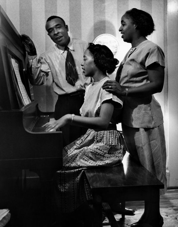 Photograph of three African American people around a piano. A woman dressed in a light colored dress plays the piano. A woman stands behind her, her hand on the piano players shoulder. A man, dressed in a white shirt and dark tie is behind them, leaning slightly to the left.