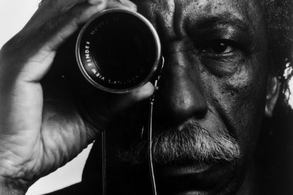 Photograph of the face of an African American man hoolding a camera lens in front of his left eye.
