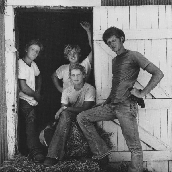 Black and white photograph of four boys in a doorway of a barn with hay on the ground and a haybale visible that one of boys is sitting on. Boys are dressed in t-shirts and jeans.