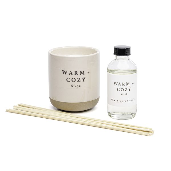 Candle in a round white jar with a rounded gold base and the words Warm + Cozy No. 32. A clear bottle with the same label and