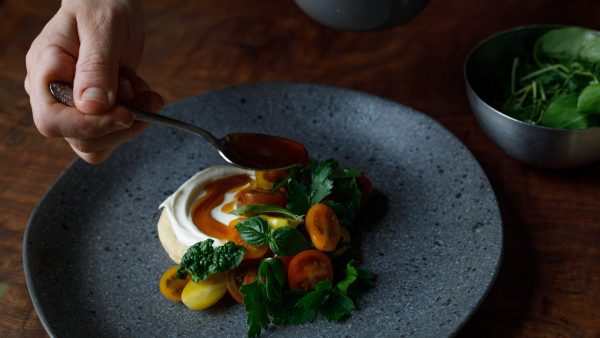 Photo of a white hand holding a spoon and drizzing sauce over a grey plate of roasted vegetables