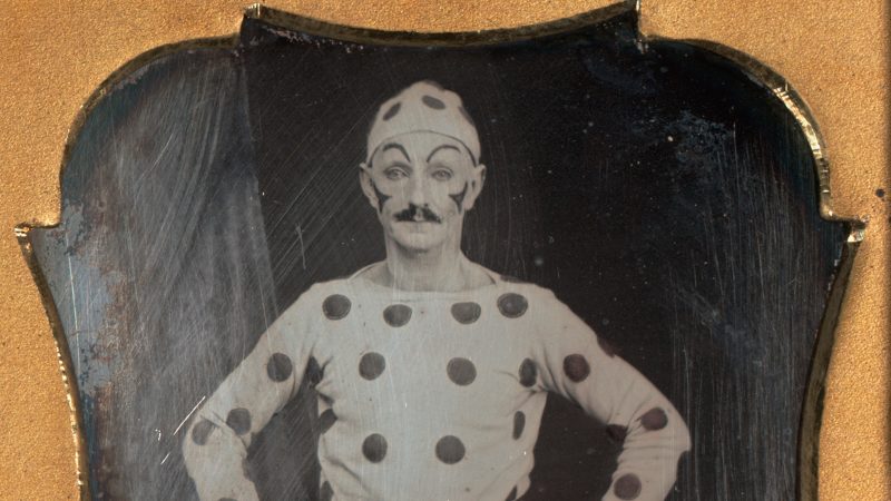 Daguerreotype of a clown standing with hands on hips, dressed in polka dots from head to toe. Frames in a light brown mat