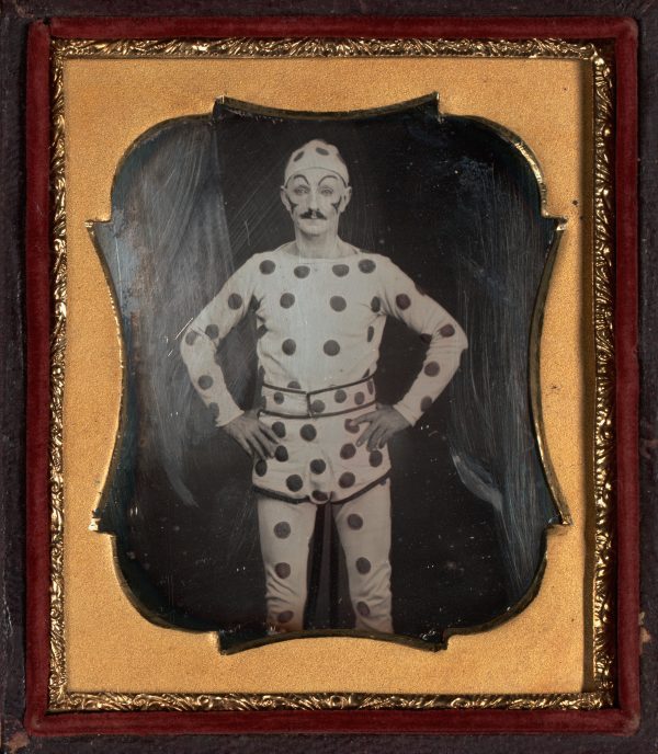 Daguerreotype of a clown standing with hands on hips, dressed in polka dots from head to toe. Frames in a light brown mat. Framed in a gold-leaf trimmed dark brown frame